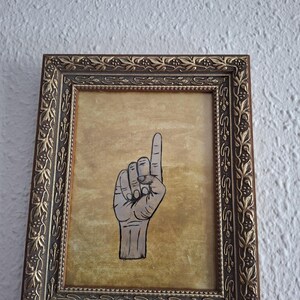 Puede incluir: Una obra de arte enmarcada que presenta una mano con el dedo índice apuntando hacia arriba. La mano está dibujada en blanco y negro sobre un fondo dorado. El marco ornamentado, de color marrón oscuro, añade una estética vintage.