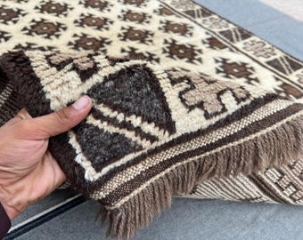 Puede incluir: Primer plano de una alfombra beige y marrón con un diseño geométrico. La alfombra tiene flecos y se levanta, revelando la parte inferior. El patrón incluye formas florales repetidas.