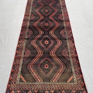 Pode incluir: Um tapete retangular de tons escuros com um padrão geométrico. O tapete apresenta um design central em forma de diamante em tons de vermelho e castanho, emoldurado por uma borda com padrões repetidos. As dimensões do tapete não são visíveis.