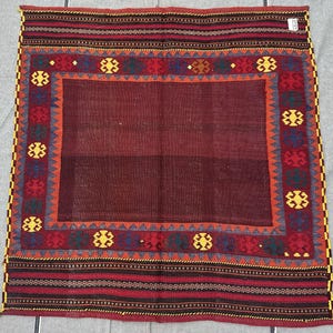 May include: A rectangular, hand-woven rug with a deep burgundy center and a patterned border. The border features a repeating floral design in red, yellow, blue, and green, framed by orange and black stripes. The rug's edges are finished with yellow fringe.