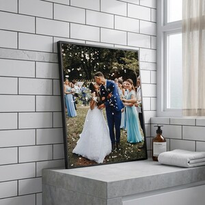 May include: A framed photograph of a wedding scene. The image features a couple kissing, surrounded by wedding guests. The bride wears a white gown, and the groom is in a blue suit. Confetti falls from above. The frame is black.