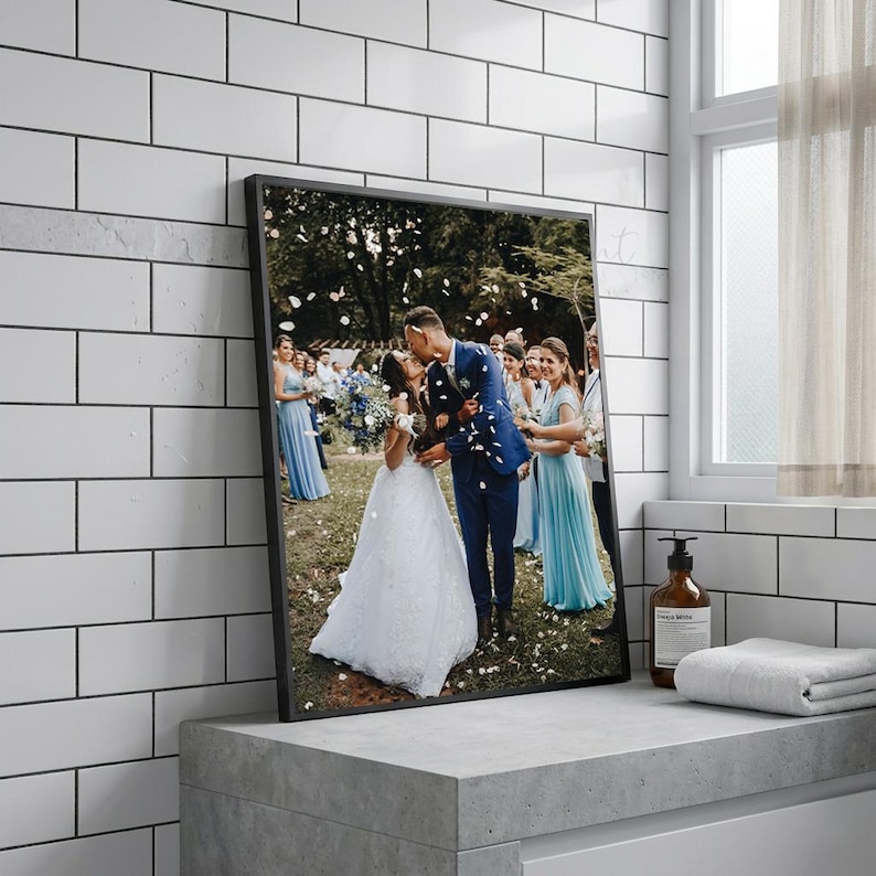 May include: A framed photograph of a wedding scene. The image features a couple kissing, surrounded by wedding guests. The bride wears a white gown, and the groom is in a blue suit. Confetti falls from above. The frame is black.