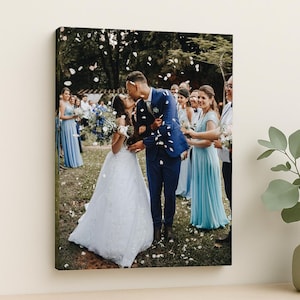 May include: A framed canvas print of a wedding scene. The image shows a couple kissing, surrounded by wedding guests. The bride wears a white gown, and the groom wears a blue suit. Guests wear blue dresses and suits. Petals fall from above.