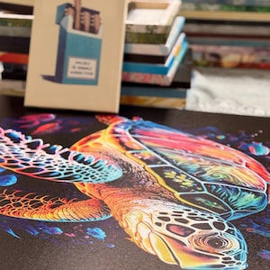 Puede incluir: Una vibrante impresi&oacute;n en lienzo que presenta una colorida tortuga marina con un patr&oacute;n de caparaz&oacute;n detallado. La obra de arte muestra un espectro de tonos, incluyendo naranja, amarillo, azul y rojo, sobre un fondo oscuro. Un libro con un dise&ntilde;o de caja de cigarrillos est&aacute; en el fondo.