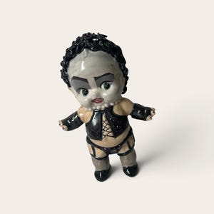 Puede incluir: Una figura de cerámica de una muñeca con piel pálida, ojos verdes y cabello oscuro rizado. La muñeca lleva un top negro estilo corsé con cordones en la parte delantera, brazaletes negros y ligueros negros sobre las piernas.