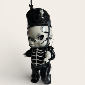 Puede incluir: Figura de cerámica de un esqueleto vestido con un uniforme negro de banda de música. La figura tiene una cara de calavera blanca con ojos negros y un sombrero negro. El uniforme del esqueleto incluye una chaqueta negra con un diseño de caja torácica blanca.
