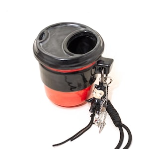 May include: A two-toned ceramic cup with a glossy black upper section and a bright red lower section. The cup features a black lid with a circular opening and a handle. A small black cat figurine charm hangs from the handle on a silver chain.