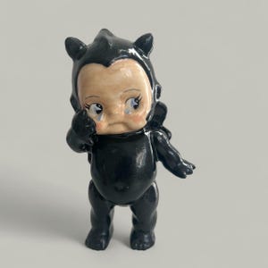 May include: A black figurine of a baby-like creature with a sad expression, tears, and small wings. The figurine has a light skin tone face with rosy cheeks and is wearing a black outfit with small ears.