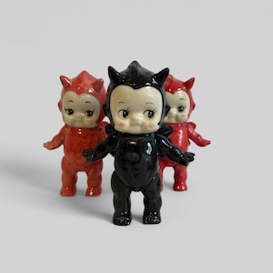 May include: Three ceramic figurines of baby devils. Two are red and one is black. They are all standing with their arms outstretched.