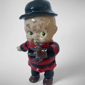 Puede incluir: Una figura de cerámica de un personaje con la cara marcada, que lleva un sombrero negro y una camisa a rayas rojas y negras. La figura tiene una mano con garras y zapatos marrones. La figura tiene ojos verdes.