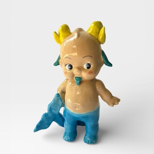 Puede incluir: Figura de cerámica de una criatura caprichosa con cuerpo bronceado, pantalones azules y cola azul. Tiene cuernos amarillos, orejas verdes y lengua turquesa. La figura tiene una estética vintage pintada a mano.