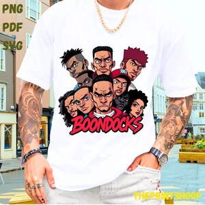 May include: White t-shirt featuring a graphic of several cartoon faces with the word "BOONDOCKS" in red. The design includes various skin tones and hairstyles. The shirt has short sleeves.