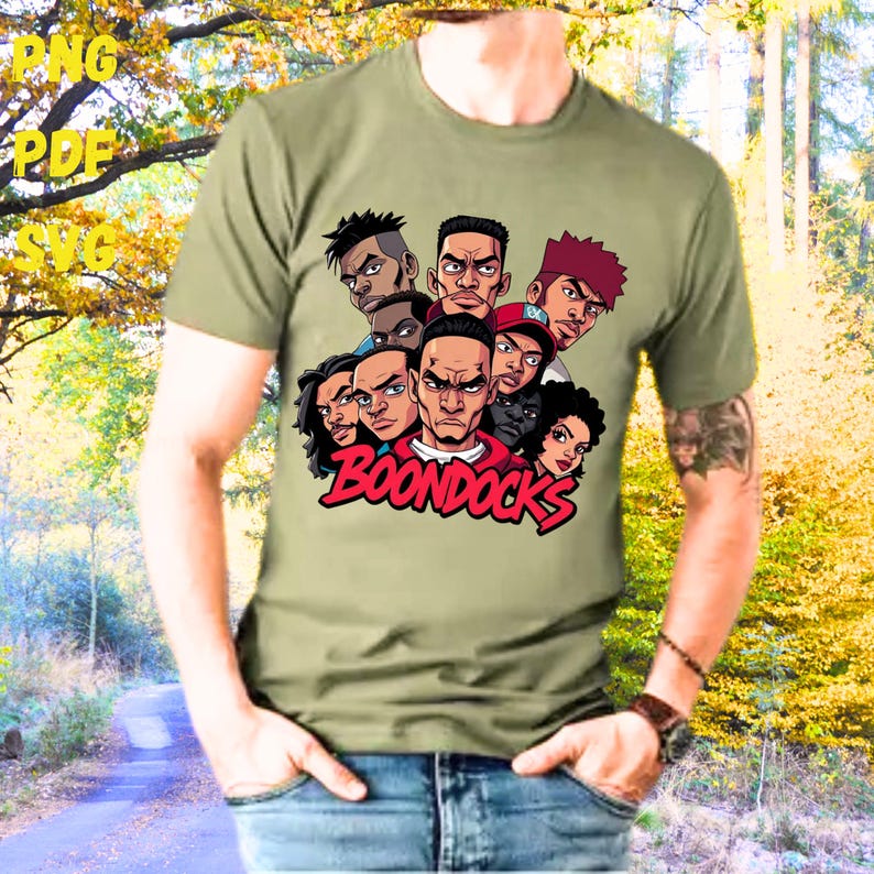 May include: Olive green t-shirt featuring a graphic of several cartoon faces with the word "BOONDOCKS" in red. The design includes various skin tones and hairstyles. The person wearing the shirt is also wearing blue jeans.