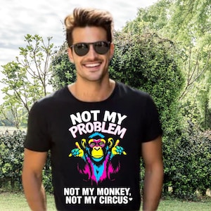 以下が含まれることがあります： 「NOT MY PROBLEM」の文字とカラフルな猿のグラフィックがプリントされた黒いTシャツ。猿は青、ピンク、黄色のアクセントで描かれています。グラフィックの下には「NOT MY MONKEY, NOT MY CIRCUS」の文字があります。