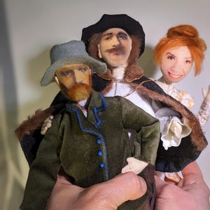 May include: Three handmade felt dolls, each with a unique face and clothing. The doll on the left wears a green jacket with blue trim and a gray hat. The doll in the middle wears a black hat and a white shirt with a brown fur collar. The doll on the right wears a white dress with a brown fur collar and a red hair bun.