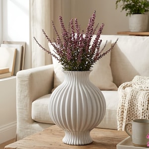 May include: A white, ribbed ceramic vase filled with purple flowers sits on a wooden table. The vase has a bulbous shape and a flared base. A neutral-toned sofa and a chunky knit blanket are in the background, creating a cozy, inviting atmosphere.