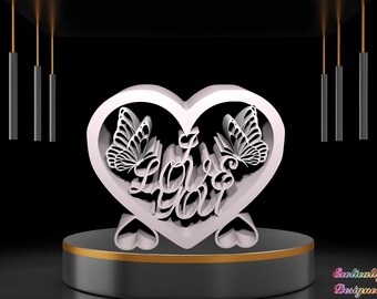 Puede incluir: Adorno blanco en forma de corazón con un diseño recortado de las palabras "Love You" y dos mariposas. El adorno está sobre un pedestal negro con un borde dorado.