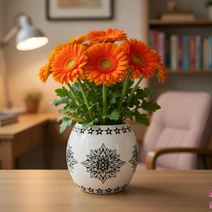 Puede incluir: Un jarrón redondo de cerámica blanca con un patrón floral negro estilo mandala y un borde de pequeñas estrellas, lleno de un vibrante ramo de gerberas de color naranja brillante con tallos y hojas verdes, sobre una superficie de madera.