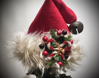 May include: A red Santa hat with a white fur trim and a decorative cluster of berries, greenery, and a wooden star. A bronze bell hangs from the side. This festive hat is a Christmas tree topper.