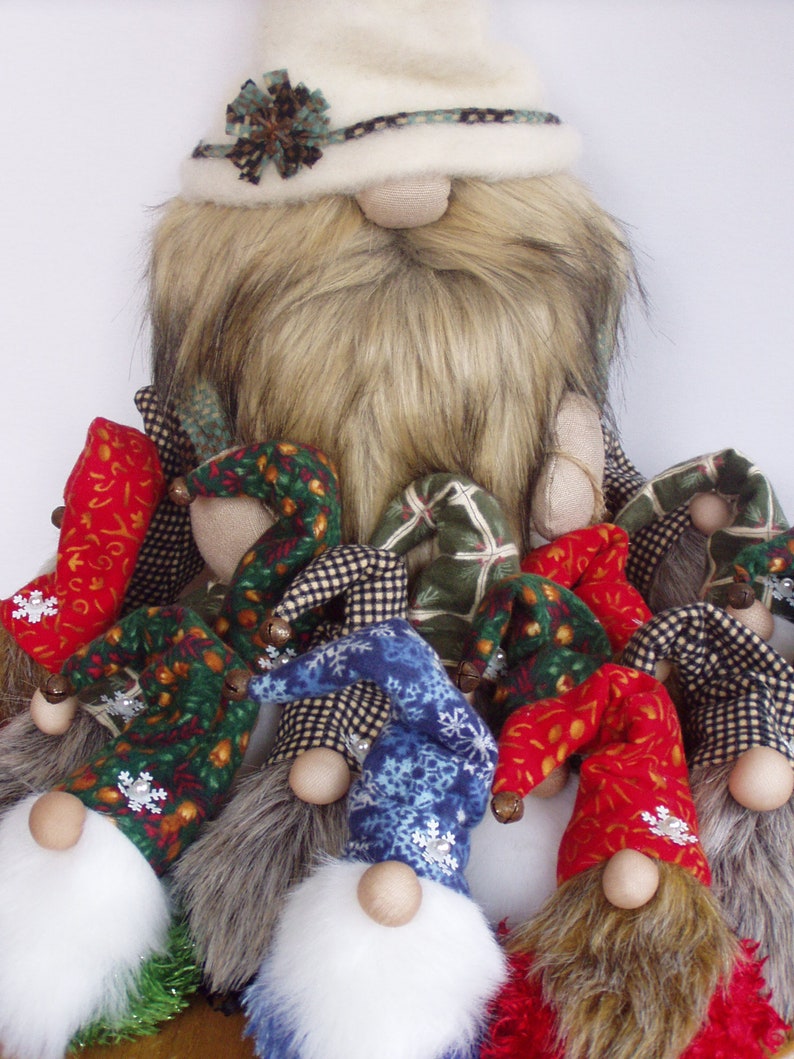 May include: A collection of handmade gnome figurines. The gnomes have long, shaggy beards, round wooden noses, and colorful, pointed hats. The hats are patterned with snowflakes, plaid, and floral designs. The main gnome has a cream-colored hat and a long, faux fur beard.