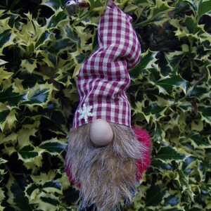 May include: A whimsical gnome ornament with a burgundy and white checkered hat and a faux fur beard. The gnome has a wooden nose and a small white snowflake on the hat. The gnome's beard is brown and the pom-pom is red.