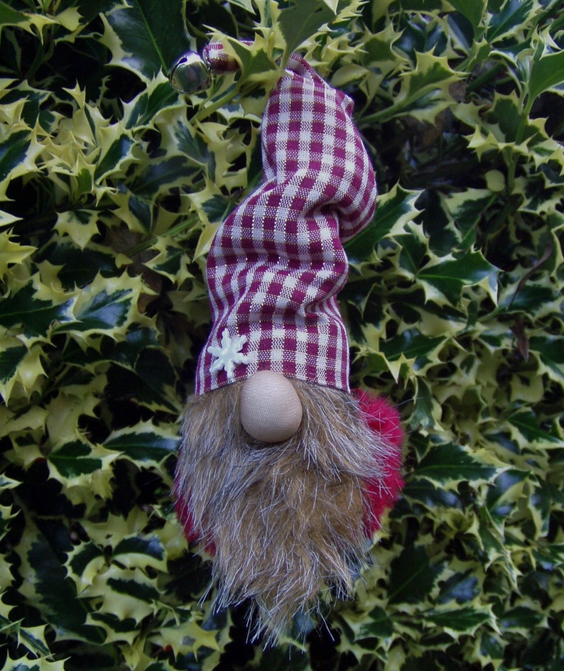 May include: A whimsical gnome ornament with a burgundy and white checkered hat and a faux fur beard. The gnome has a wooden nose and a small white snowflake on the hat. The gnome's beard is brown and the pom-pom is red.
