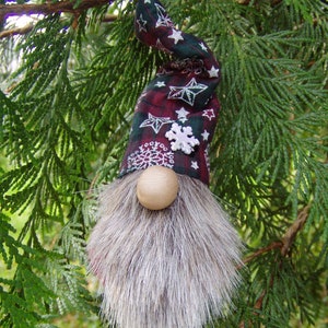 May include: A whimsical Christmas gnome ornament with a long gray faux fur beard and a round wooden nose. The gnome's hat is burgundy and green plaid with white star and snowflake accents. The ornament hangs from a green tree branch.