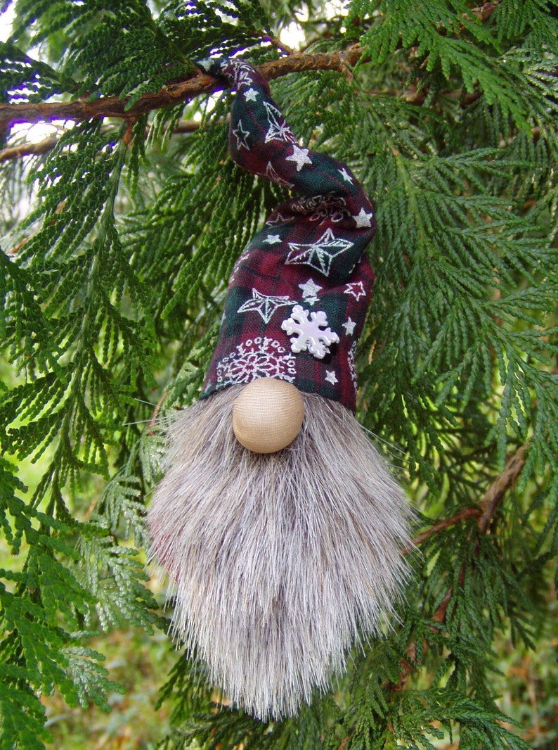 May include: A whimsical Christmas gnome ornament with a long gray faux fur beard and a round wooden nose. The gnome's hat is burgundy and green plaid with white star and snowflake accents. The ornament hangs from a green tree branch.