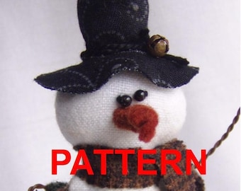 May include: A fabric snowman figurine wearing a black patterned top hat with a small brass bell, a brown textured scarf, and a dark green base. The snowman has black bead eyes and a rust-colored nose. The text PATTERN is visible in red.
