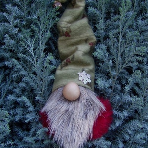 May include: A whimsical Christmas gnome with a long, olive green hat adorned with red berry accents and a silver snowflake. The gnome features a light brown wooden nose, a fluffy gray beard, and a red pom-pom base. A small bell hangs from the hat's tip.