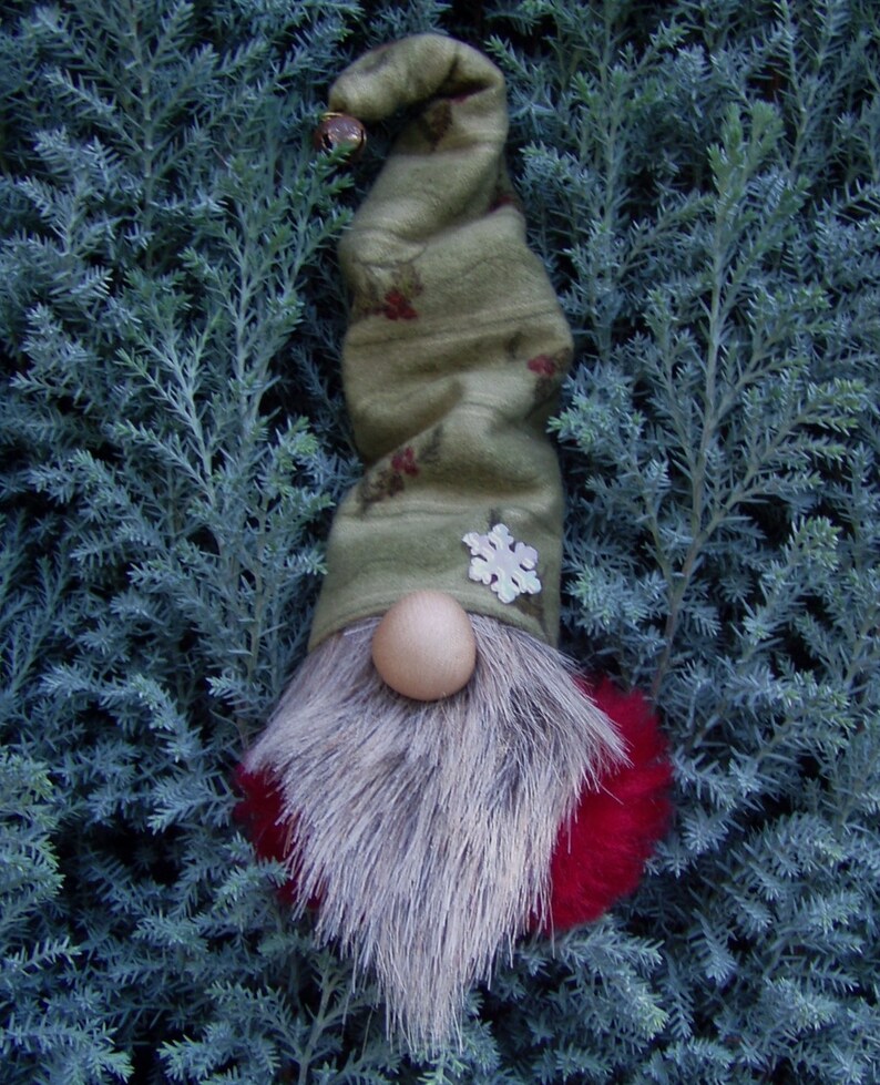 May include: A whimsical Christmas gnome with a long, olive green hat adorned with red berry accents and a silver snowflake. The gnome features a light brown wooden nose, a fluffy gray beard, and a red pom-pom base. A small bell hangs from the hat's tip.