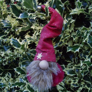 May include: A festive gnome ornament with a red hat adorned with gold dots and a white snowflake. The gnome has a wooden nose and a fluffy, gray beard. A small silver bell hangs from the hat. The ornament is set against a backdrop of green and yellow holly leaves.