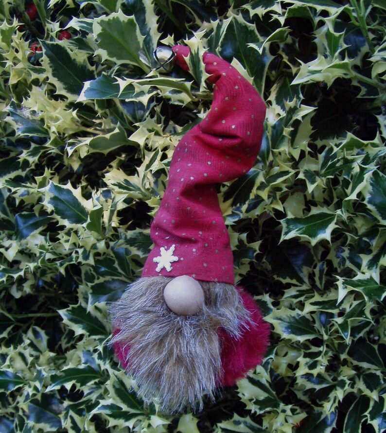 May include: A festive gnome ornament with a red hat adorned with gold dots and a white snowflake. The gnome has a wooden nose and a fluffy, gray beard. A small silver bell hangs from the hat. The ornament is set against a backdrop of green and yellow holly leaves.