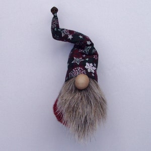May include: A whimsical gnome figurine with a long, gray faux fur beard and a wooden nose. The hat is patterned with white stars and snowflakes on a dark red and black plaid background. The hat has a curved shape and a small brown knob at the tip.