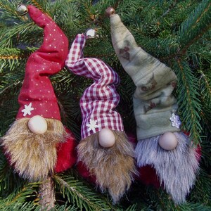 May include: Three whimsical Christmas gnome ornaments with long, twisted hats and faux fur beards. The hats are red with white polka dots, red and white gingham, and green with floral patterns. Each gnome has a wooden nose and a snowflake accent.