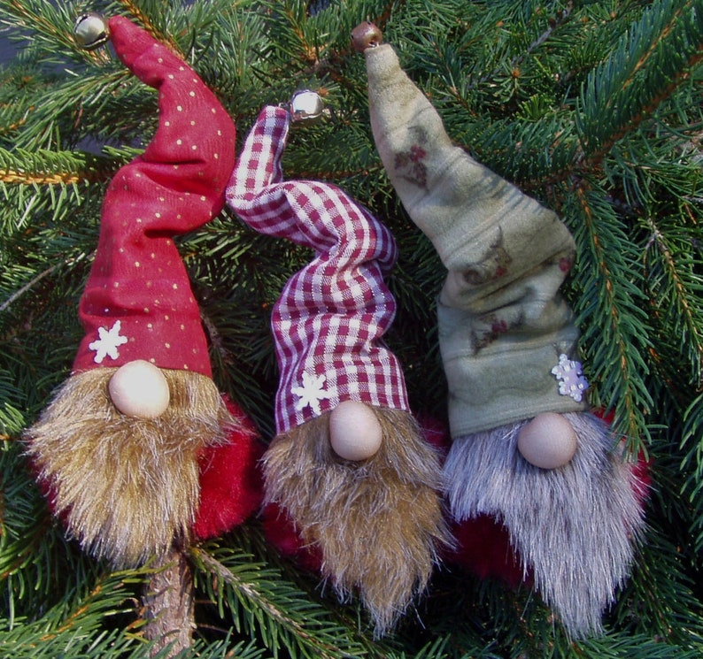 May include: Three whimsical Christmas gnome ornaments with long, twisted hats and faux fur beards. The hats are red with white polka dots, red and white gingham, and green with floral patterns. Each gnome has a wooden nose and a snowflake accent.