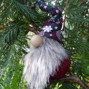 May include: A handmade Christmas gnome ornament with a burgundy body and a long, silver-gray faux fur beard. The gnome's hat is patterned with white snowflakes and stars on a dark green and burgundy background. A wooden ball serves as the nose.