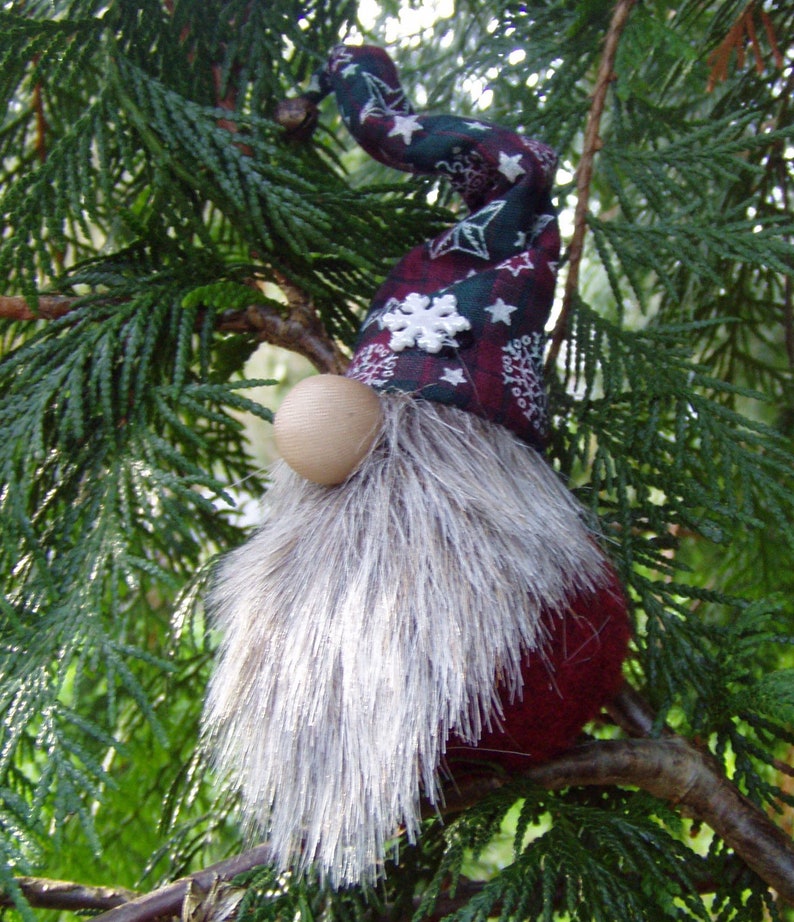 May include: A handmade Christmas gnome ornament with a burgundy body and a long, silver-gray faux fur beard. The gnome's hat is patterned with white snowflakes and stars on a dark green and burgundy background. A wooden ball serves as the nose.