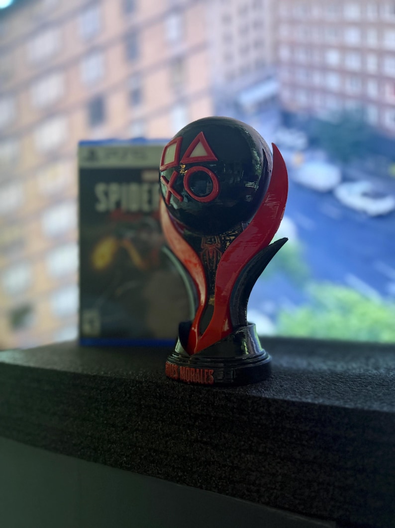 Puede incluir: Un trofeo negro y rojo con los s&iacute;mbolos de PlayStation y la inscripci&oacute;n "Miles Morales" en la base. En el fondo, se ve una caja del videojuego "Spider-Man Miles Morales".