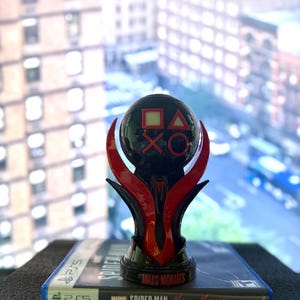 Puede incluir: Un trofeo negro y rojo de PlayStation 5 con los s&iacute;mbolos de PlayStation en una esfera. El trofeo est&aacute; encima de una caja de juego de Marvel Spider-Man: Miles Morales para PS5. El fondo muestra una vista de la ciudad.