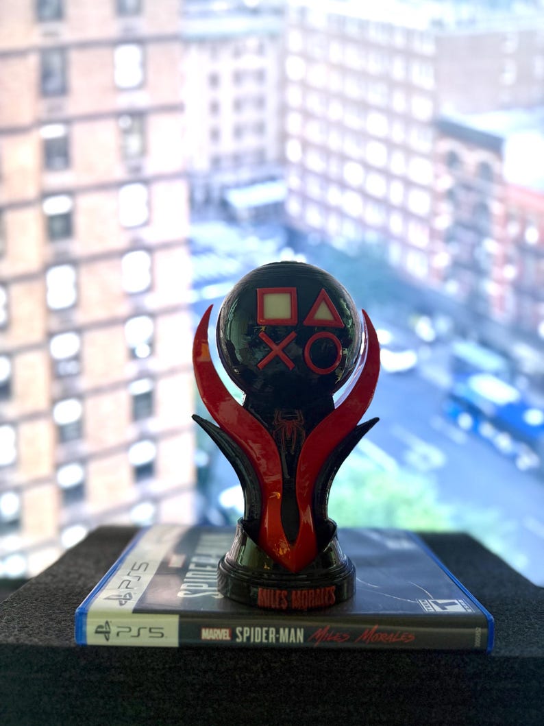 Puede incluir: Un trofeo negro y rojo de PlayStation 5 con los s&iacute;mbolos de PlayStation en una esfera. El trofeo est&aacute; encima de una caja de juego de Marvel Spider-Man: Miles Morales para PS5. El fondo muestra una vista de la ciudad.