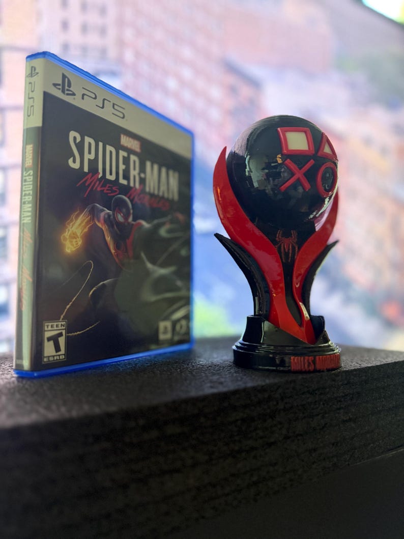 Puede incluir: Una caja de juego de PS5 para Marvel's Spider-Man: Miles Morales junto a un trofeo negro y rojo. El trofeo tiene los s&iacute;mbolos de PlayStation en la parte superior y las palabras "Miles Morales" en la base.