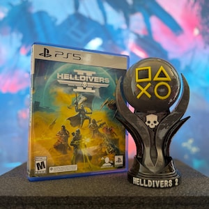 Puede incluir: Una caja de juego de PS5 para "HELLDIVERS" junto a un trofeo negro y gris. El trofeo tiene una calavera y los símbolos de PlayStation en amarillo. La caja del juego tiene un borde azul y una ilustración de ciencia ficción.
