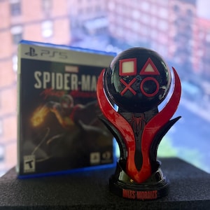 Puede incluir: Un trofeo negro y rojo con s&iacute;mbolos de mando de PlayStation y las palabras "Miles Morales". Una caja de juego de PS5 con "Spider-Man: Miles Morales" est&aacute; en el fondo.