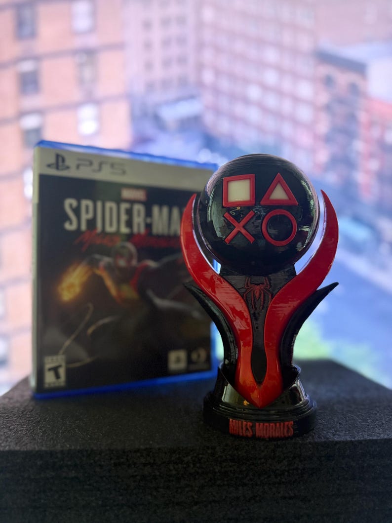 Puede incluir: Un trofeo negro y rojo con s&iacute;mbolos de mando de PlayStation y las palabras "Miles Morales". Una caja de juego de PS5 con "Spider-Man: Miles Morales" est&aacute; en el fondo.