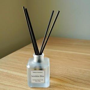 May include: A frosted glass diffuser bottle with black reeds. The label reads "Sunshine Bliss" with scent notes. The bottle is on a wooden surface. The product is 4 fl oz / 120ml.