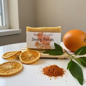 May include: A bar of "Zesty Patch" soap with orange and patchouli, accompanied by dried orange slices, an orange, and a pile of orange powder. The soap is labeled "Vedanta Aromatics" and weighs 4oz.