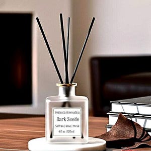 May include: A clear glass diffuser bottle with black reeds, labeled "Dark Suede" with scent notes. The bottle sits on a white marble coaster. A piece of dark brown suede is next to the bottle.