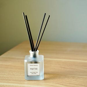 May include: A frosted glass diffuser bottle with black reeds. The label reads "High Tide". The bottle sits on a light wood surface. The background is a neutral color.