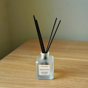 May include: A frosted glass diffuser bottle with black reeds. The label reads "Beach Linen" with the brand name "Vedanta Aromatics". The bottle sits on a light brown wooden surface, with a neutral background.